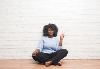 Young african american woman sitting on the floor at home smiling with happy face winking at the camera doing victory sign. Number two.