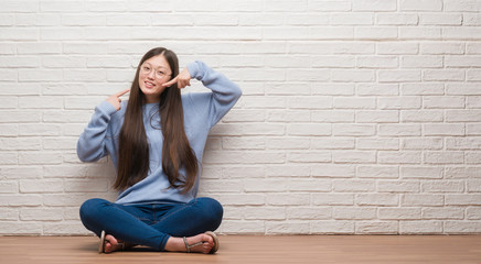 Fototapeta na wymiar Young Chinese woman sitting on the floor over brick wall smiling confident showing and pointing with fingers teeth and mouth. Health concept.