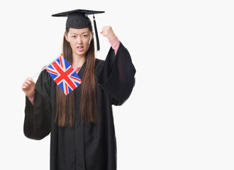 Young Chinese woman wearing graduate uniform holding United kingdom flag annoyed and frustrated shouting with anger, crazy and yelling with raised hand, anger concept