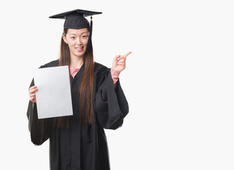 Young Chinese woman wearing graduate uniform holding paper degree very happy pointing with hand and finger to the side