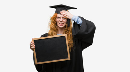 Young redhead student woman wearing graduated uniform holding blackboard stressed with hand on...