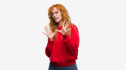 Young redhead woman wearing red sweater afraid and terrified with fear expression stop gesture with hands, shouting in shock. Panic concept.