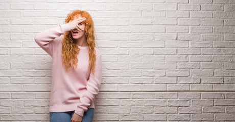 Young redhead woman standing over brick wall peeking in shock covering face and eyes with hand, looking through fingers with embarrassed expression.