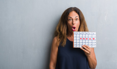Middle age hispanic woman standing over grey grunge wall holding period calendar scared in shock...