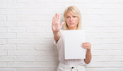 Adult caucasian woman over white brick wall holding blank paper sheet with open hand doing stop...