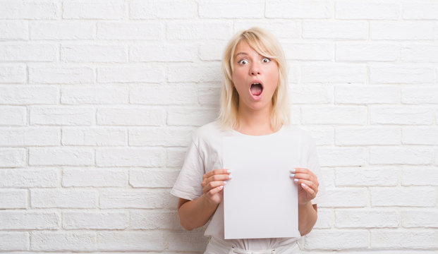 Adult caucasian woman over white brick wall holding blank paper sheet scared in shock with a surprise face, afraid and excited with fear expression