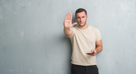 Young caucasian man over grey grunge wall texting a message using smartphone with open hand doing stop sign with serious and confident expression, defense gesture