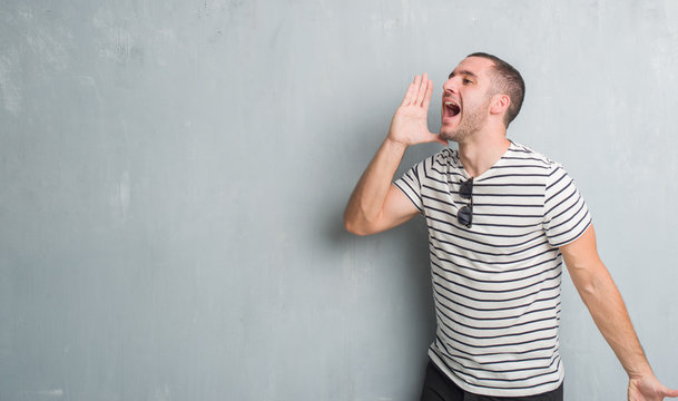 Young caucasian man over grey grunge wall shouting and screaming loud to side with hand on mouth. Communication concept.