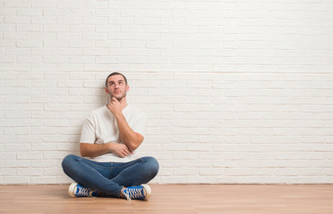 Young caucasian man sitting on the floor over white brick wall with hand on chin thinking about...