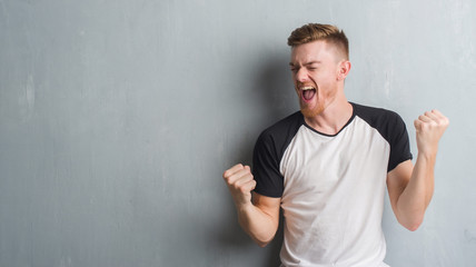Young redhead man over grey grunge wall very happy and excited doing winner gesture with arms raised, smiling and screaming for success. Celebration concept.