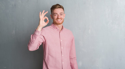 Young redhead man over grey grunge wall wearing pink shirt smiling positive doing ok sign with hand and fingers. Successful expression.