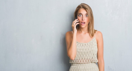Beautiful young woman over grunge grey wall speaking on the phone scared in shock with a surprise...