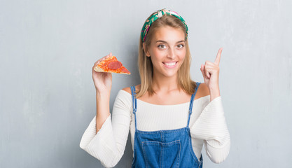 Beautiful young woman over grunge grey wall eating pepperoni pizza slice surprised with an idea or question pointing finger with happy face, number one