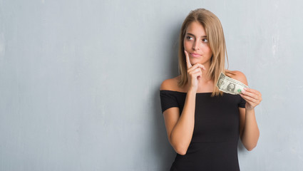 Beautiful young woman over grunge grey wall holding a dollar serious face thinking about question,...