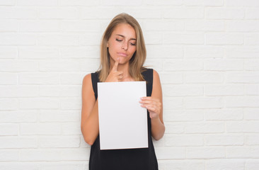 Beautiful young woman over white brick wall holding blank paper sheet serious face thinking about...