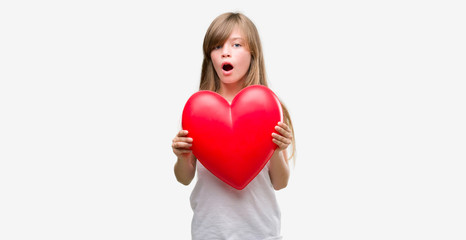 Young blonde toddler holding a red heart scared in shock with a surprise face, afraid and excited with fear expression