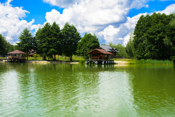 Fototapeta premium A beautiful image of a landscape from center of a river surrounded by trees and reeds on the shore against a blue sky in the clouds. Wooden gazebo on the beach. Reflection, water, tourist destination