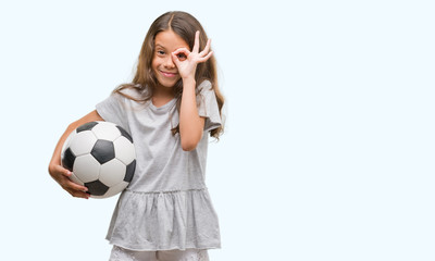 Brunette hispanic girl holding soccer football ball with happy face smiling doing ok sign with hand on eye looking through fingers