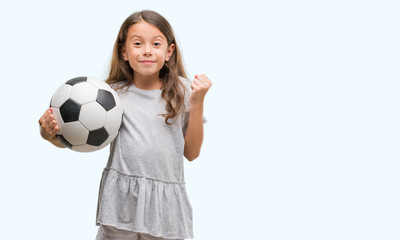 Brunette hispanic girl holding soccer football ball screaming proud and celebrating victory and success very excited, cheering emotion