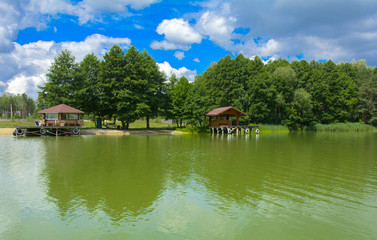 A beautiful image of a landscape from the center of a river surrounded by trees and reeds on the shore against a blue sky in the clouds. Wooden gazebo on beach. Reflection, water, tourist destination