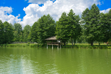 Fototapeta na wymiar A beautiful image of a landscape from center of a river surrounded by trees and reeds on the shore against a blue sky in the clouds. Wooden gazebo on the beach. Reflection, water, tourist destination