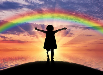 Fototapeta premium A happy child is a disabled girl with a prosthetic leg in the open air on the background of sea sunset and rainbow