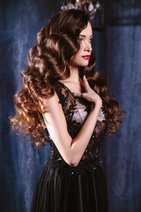 Fototapeta na wymiar Young woman with long curly hair and makeup in evening long luxury dress, posing in a dark interior room. fashion beauty portrait