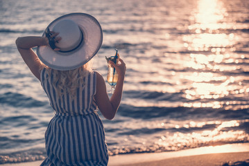 Girl wearing striped dress walking standing and holding a hat and a cocktail on the sea shore. Marine clothing style.