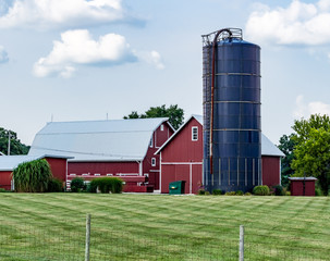 Modern, bright red barn, farm buildings and blue silo, with blue sky and puffy clouds. Working family farm with beautiful barns and lawn © Kimberly Boyles