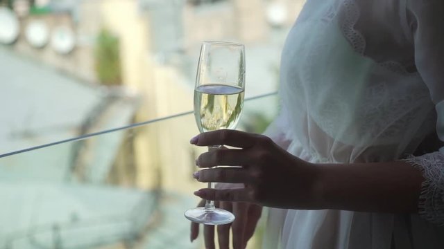 Young woman on balcony or terrace in white lingerie - bra and bathrobe drinking champagne from glass. City view on background slowmotion