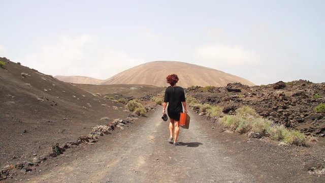 Woman with vintage travel case walks along a straight volcanic road towards a volcano in the distance