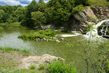 The waterfall on the river flows through and over the rocks covered with lichen and moss against a background of green vegetation and a blue sky.