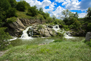 The waterfall on the river flows through and over the rocks covered with lichen and moss against a background of green vegetation and a blue sky.