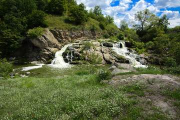 The waterfall on the river flows through and over the rocks covered with lichen and moss against a background of green vegetation and a blue sky.