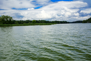 A beautiful image of landscape from the center of the river, surrounded by trees and reeds on the shore and distant horizon against the blue sky in clouds. Reflection, water, tourist destination