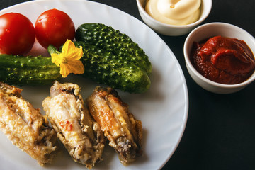 Baked chicken wings, mayonnaise, tomato paste and fresh organic vegetables on a white plate on a black background. Dietary healthy food for dinner. Copy space