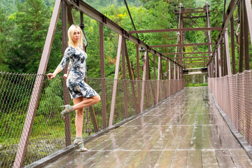 the blonde girl stands on a bridge constructed of metal and wood against the backdrop of a green forest and mountains, taking a pose by lifting her leg, bending and leaning her elbows on the railing