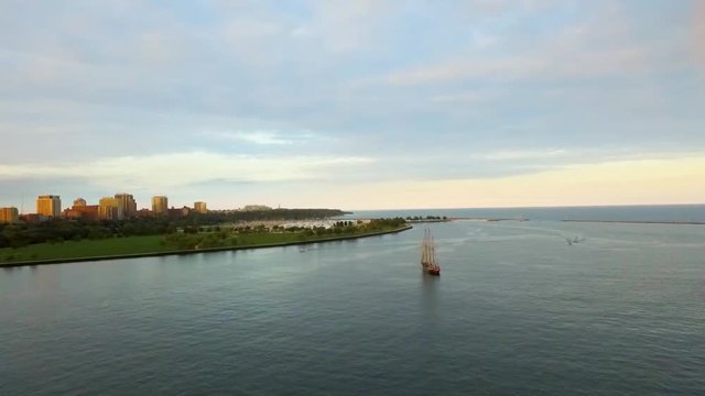 A stunning aerial drone clip flies over the gorgeous harbor and lakefront of Milwaukee Wisconsin as a tall ship and two boats navigate calm waters at sunset. A perfect establishing shot.