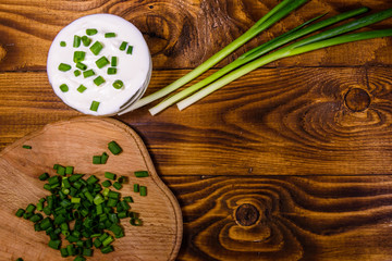 Glass bowl with sour cream and cutting board with chopped green onion on wooden table. Top view