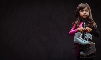 Fototapeta na wymiar Lonely little girl dressed in sportswear holds ice skates. Isolated on dark textured background.