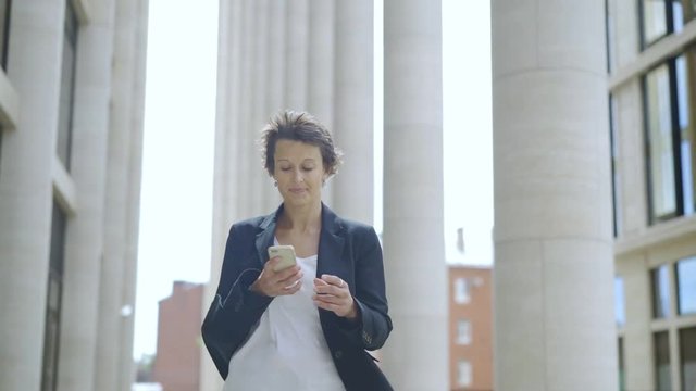 Slow motion of determined middle-adult woman texting on her phone while walking confidently down street
