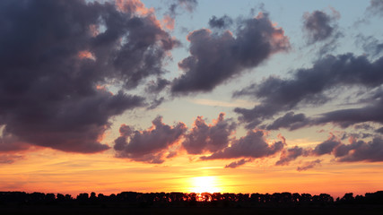The sunset of orange, red, yellow, magenta colors is in a fat country. Dark purple clouds are above on horizon and sunset. Some clouds reflect the sunlight making bright border around them.