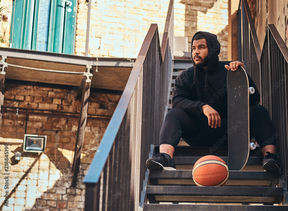 Wall mural Dark skinned bearded hipster guy dressed in a black hoodie and sports shorts holds a skateboard while sitting on stairs in ghetto.