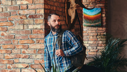 Fototapeta na wymiar Handsome bearded hipster male in a blue fleece shirt and jeans with backpack leaning against a brick wall at a studio with loft interior.