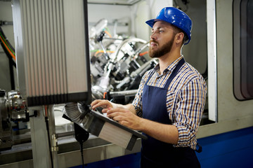 Side view portrait of machine operator wearing hardhat standing at control panel pressing buttons,...