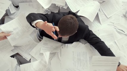 A young tired man is sitting in a pile of papers, signing many documents. An office employee can not cope with work, looks at the clock.