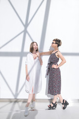 Fototapeta premium Two fashionable girlfriends stand in full swing at the background of a white wall with shadows. Portrait of two stylish attractive girls on the background of a white wall.