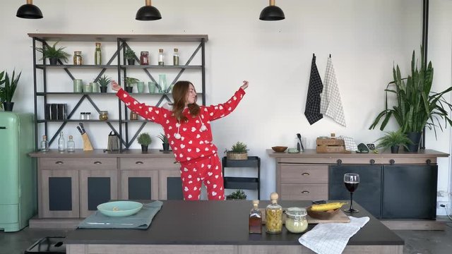 Adorable young woman in funny pajamas with hears dancing and twisting around in kitchen behind table, cheerful and happy