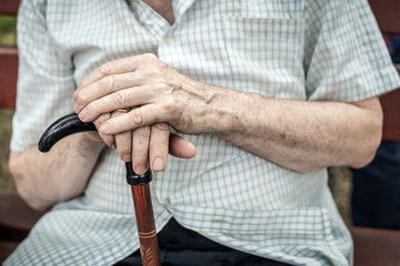 Senior person sitting on wooden bench outdoors. Old man hands holding walking stick. Poverty, loneliness and hopelessness concept