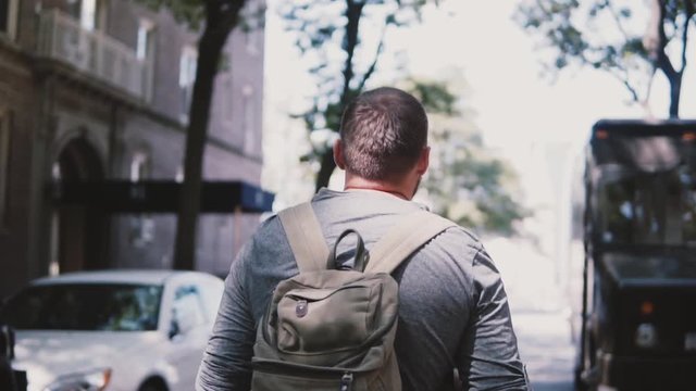 Camera follows young relaxed male freelance worker with backpack walking along shady street in Brooklyn slow motion.